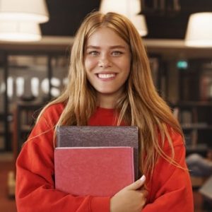 cheerful-smiling-readhead-girl-holding-books-standing-library-bookstore-min